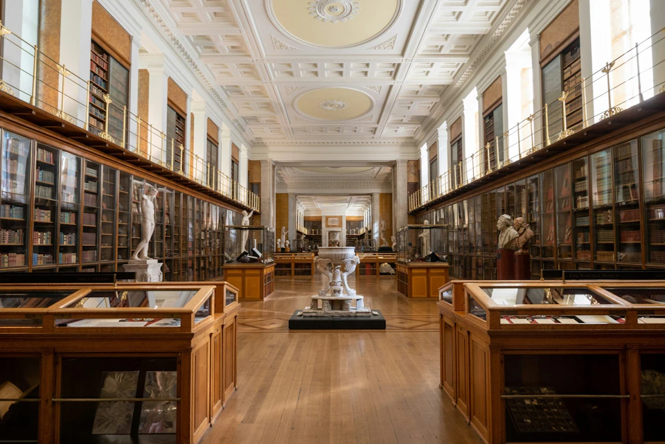 A spacious museum gallery with glass display cases, sculptures, and ornate ceilings.