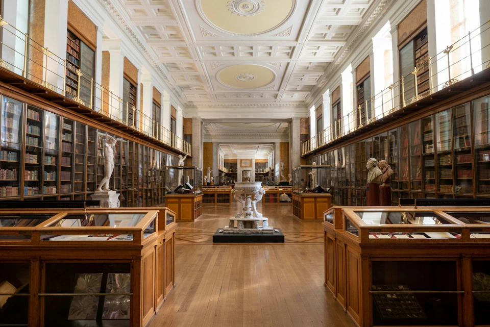 A spacious museum gallery with glass display cases, sculptures, and ornate ceilings.