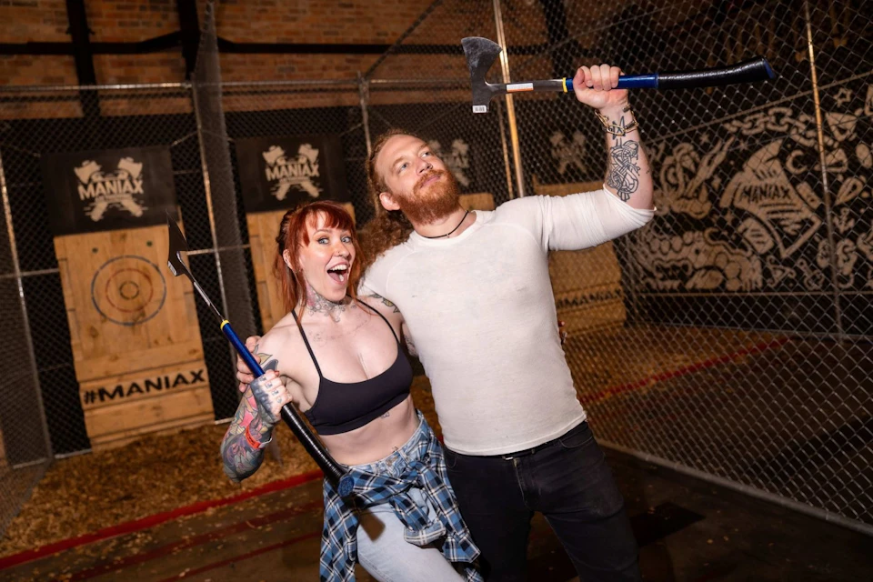 Two people smiling and posing with axes in an indoor axe-throwing venue, with target boards visible in the background.