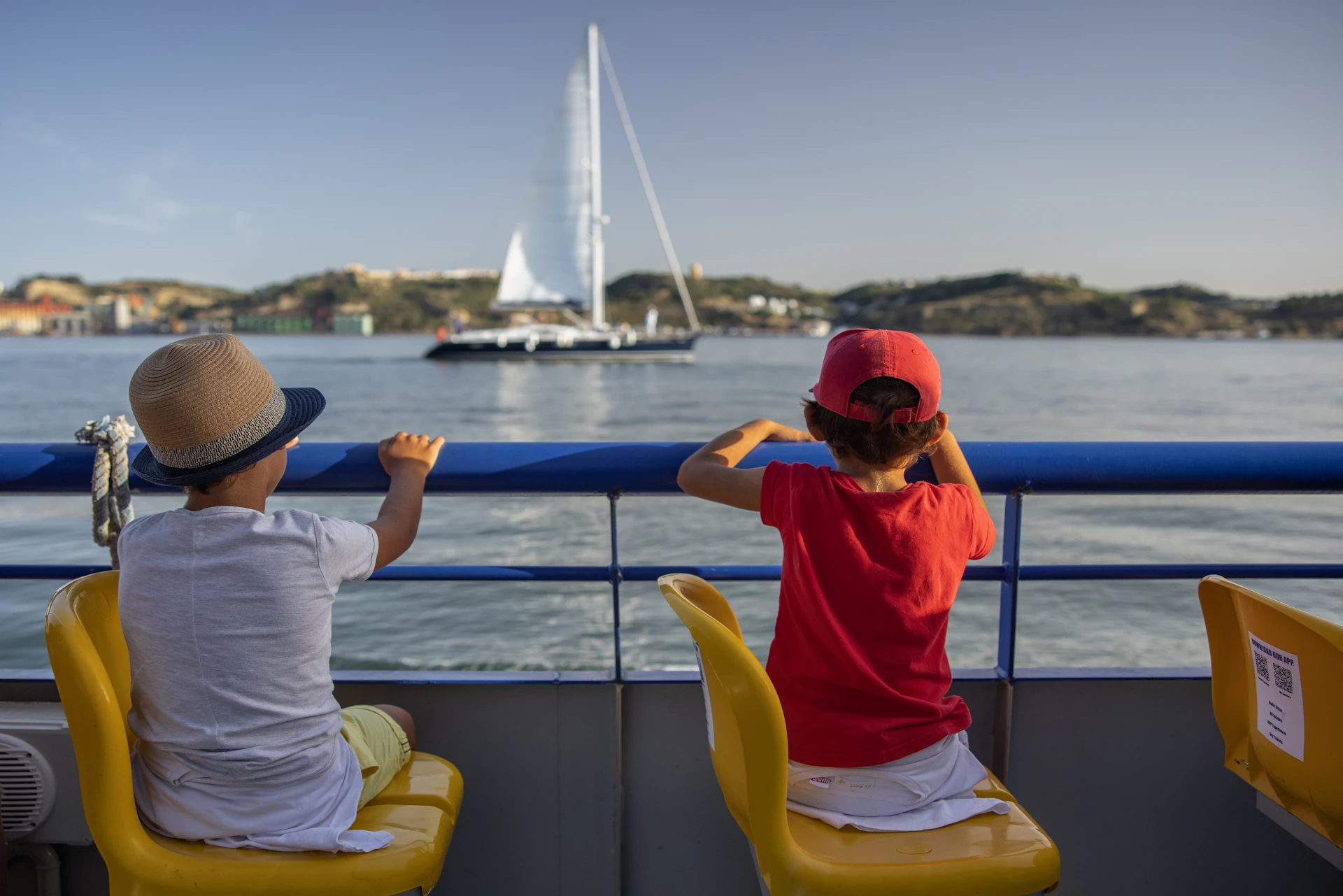Two children sit on a boat, looking at a distant sailboat on calm water with hills in the background.