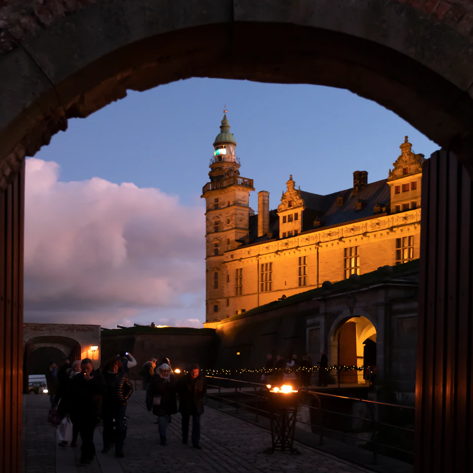 People walking through an archway toward a lit-up historic building with a clock tower at dusk.