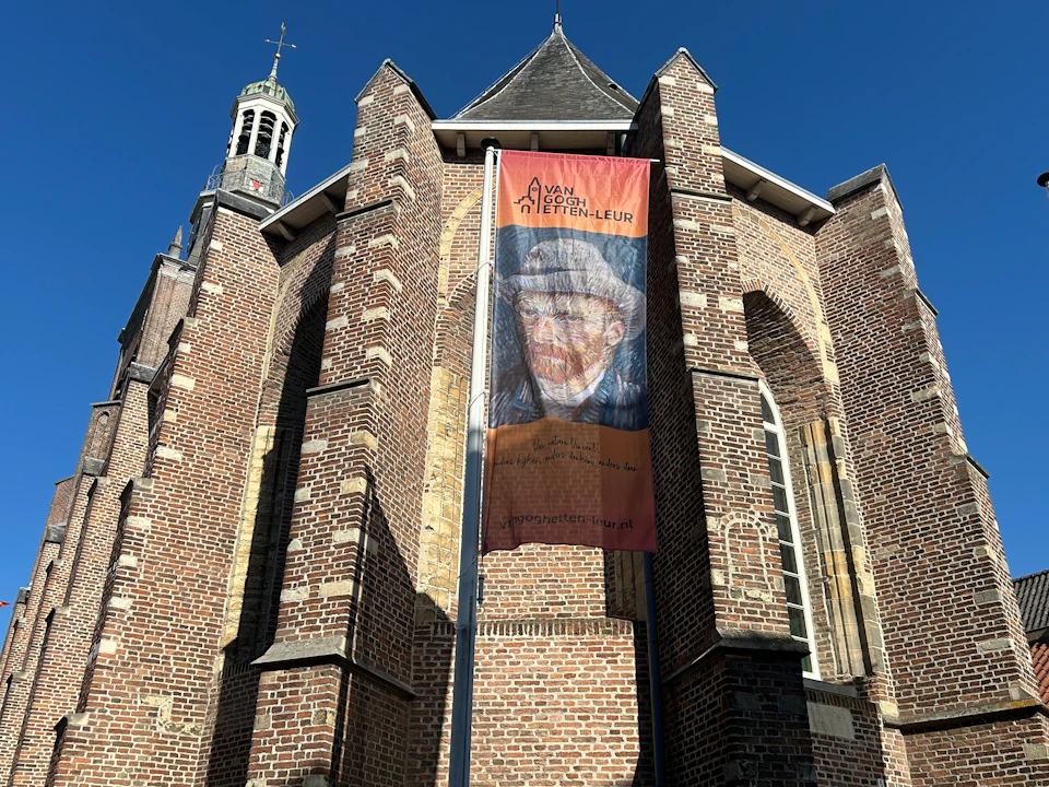 Tall brick church with arched windows and a vertical banner depicting Van Gogh's face, against a clear blue sky.