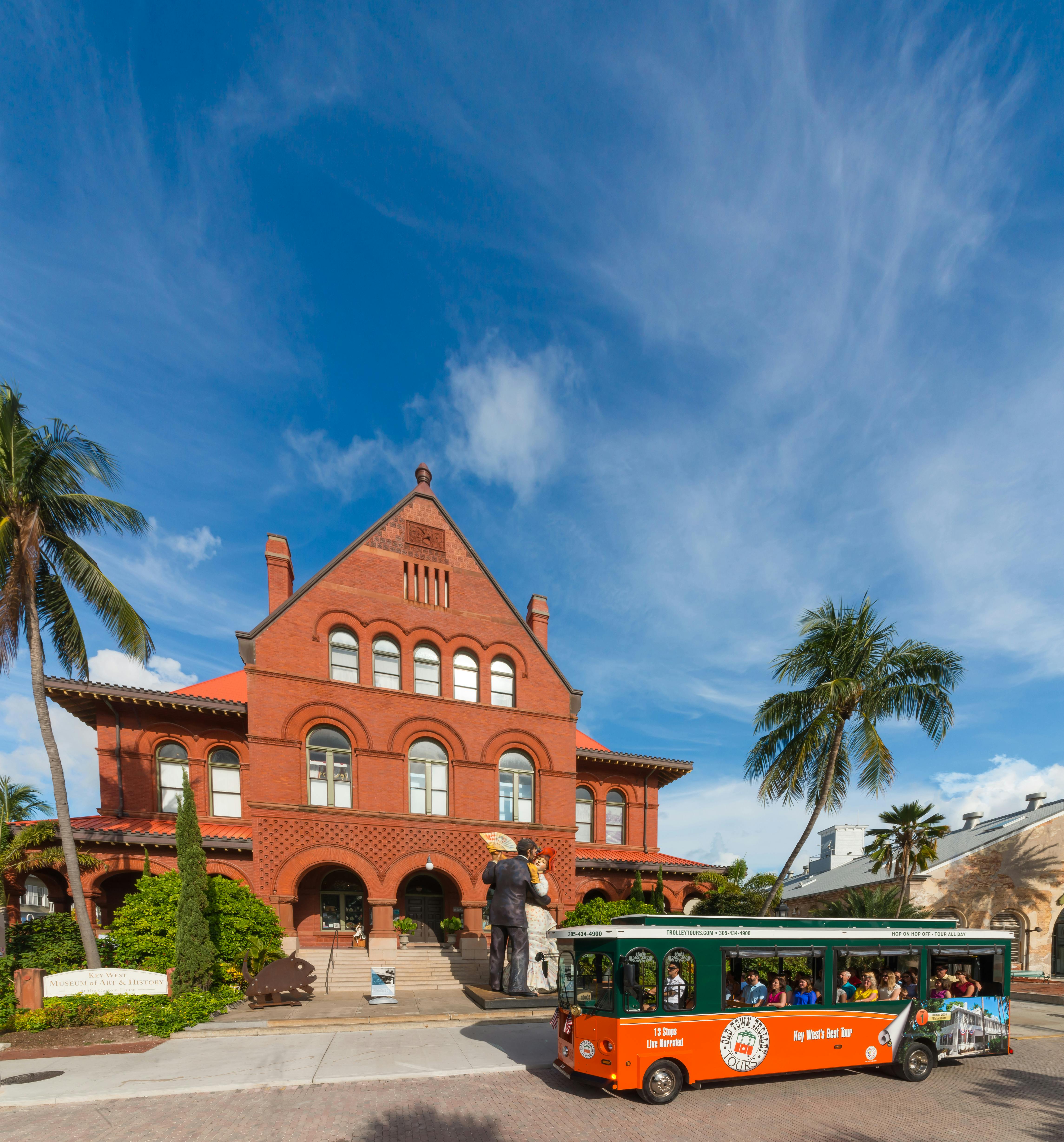Jasně červená cihlová budova s klenutými okny, palmami a zeleným trolejbusem před sebou za jasného slunečného dne.