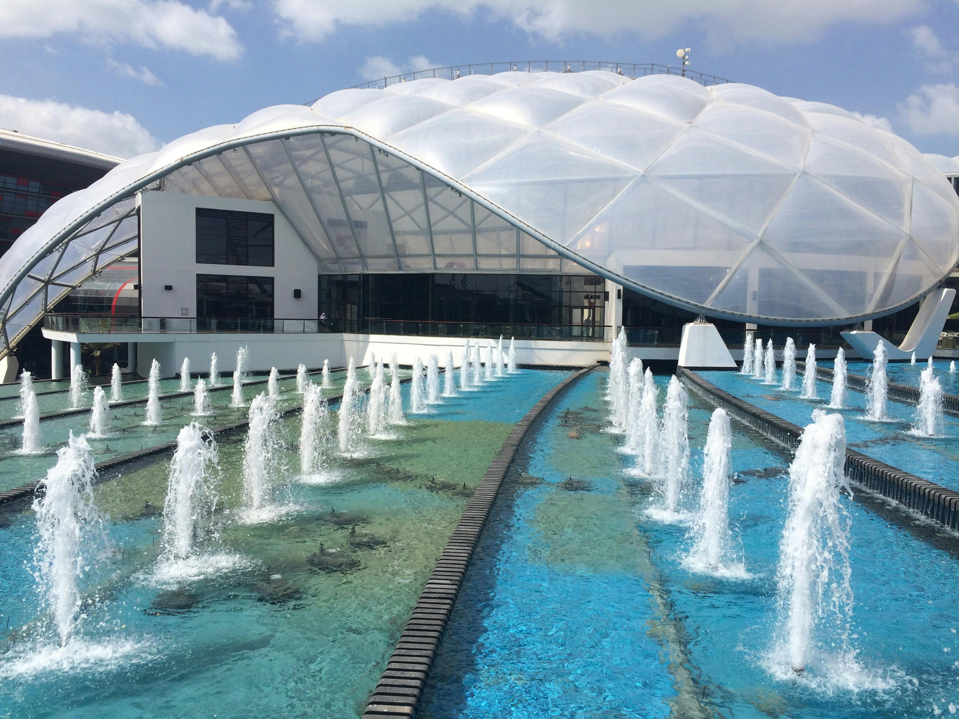 A modern building with a large, dome-shaped translucent roof and several water fountains in front.