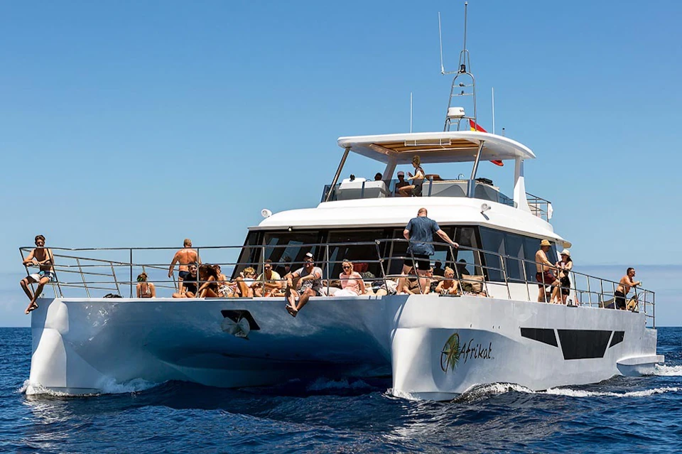 A group of people relaxing and sunbathing on the deck of a white yacht named "Afrikat," sailing on the blue ocean under a clear sky.