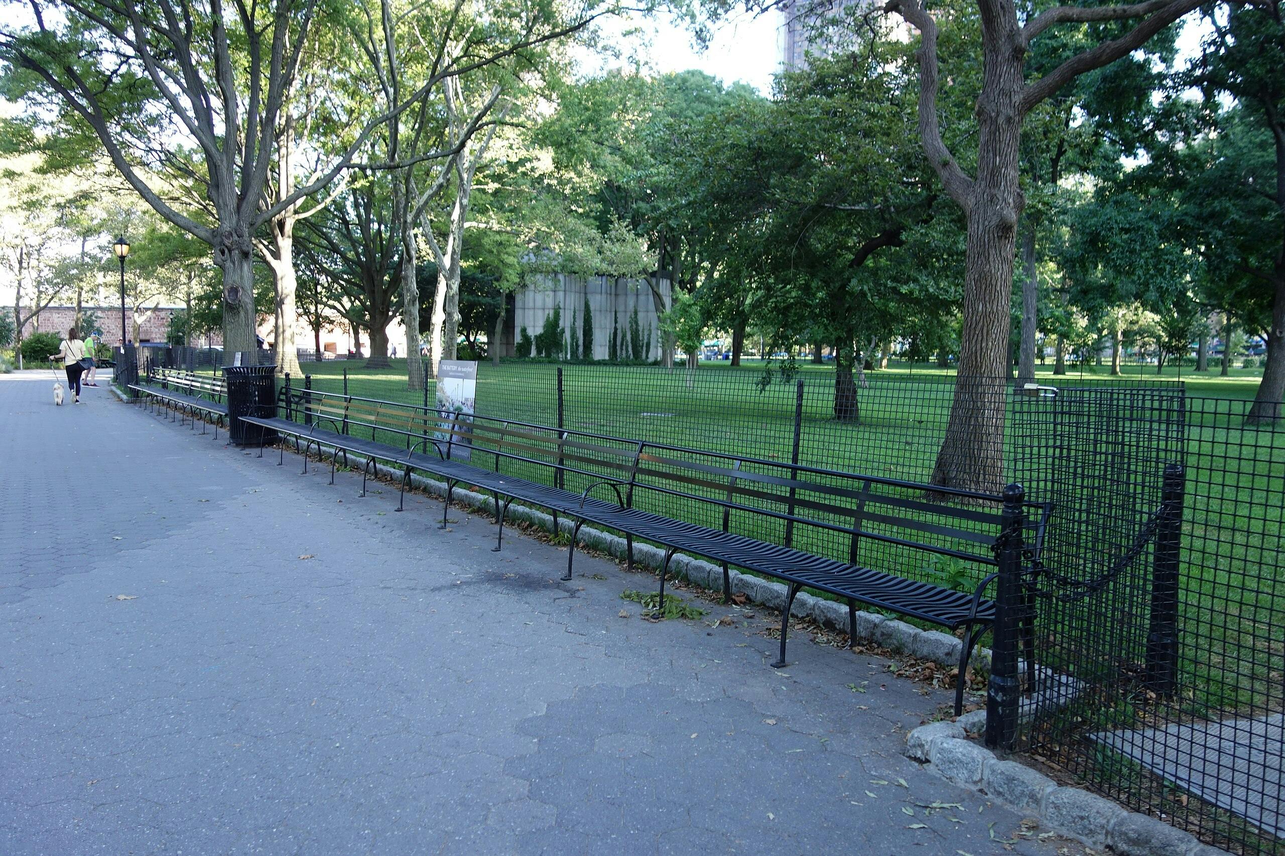 人们在空荡荡的公园长椅，高大的树木和围栏绿色草坪旁边的小路上遛狗。