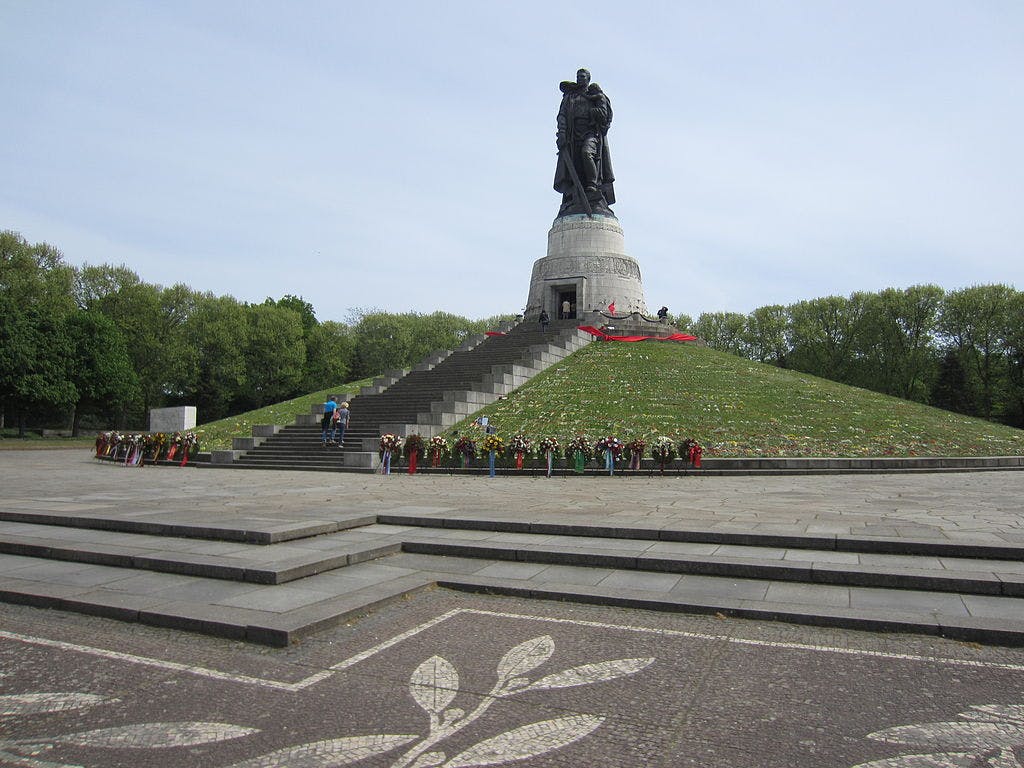 A large statue atop a hill with steps, surrounded by flowers and trees. Two people are ascending the steps.