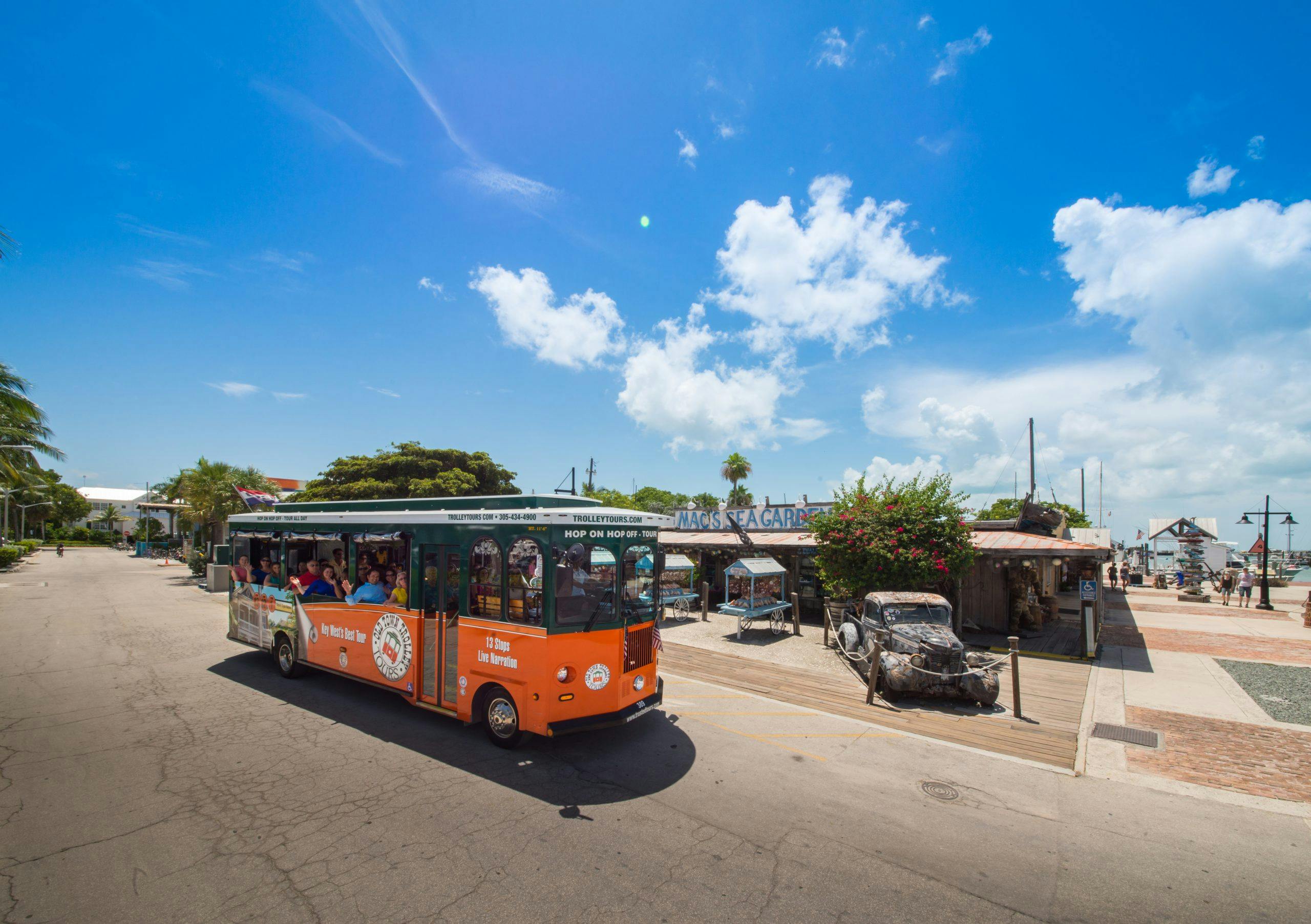 Oranžový a zelený vozík s cestujícími na slunné ulici. V pozadí jsou budovy, rustikální auto a jasná obloha s mraky.