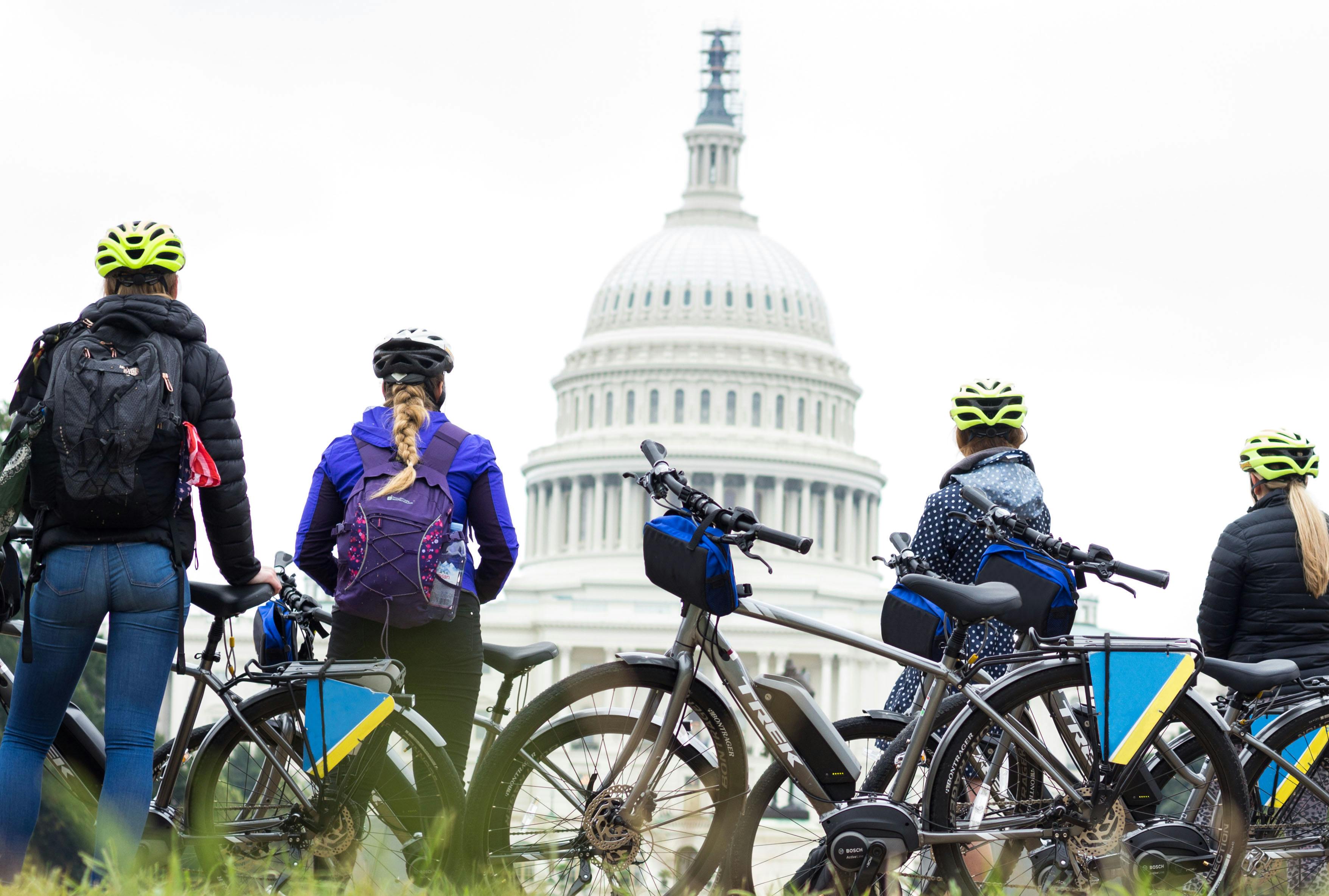 Three cyclists with helmets and backpacks stand next to their bikes, facing the US Capitol building.