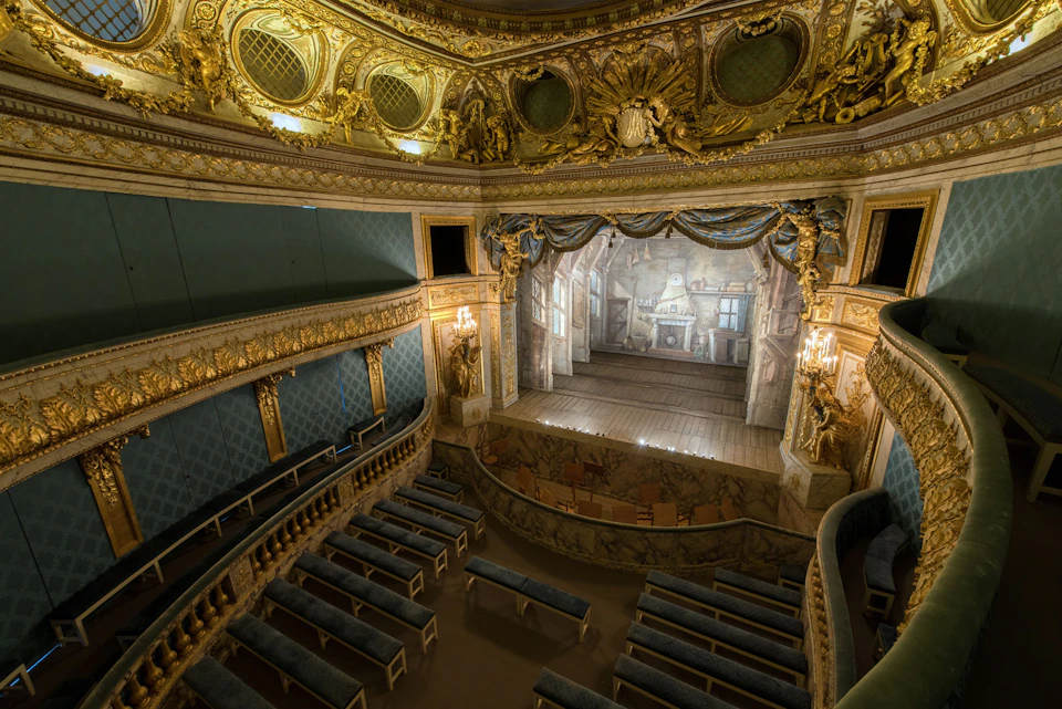 Ornate, empty theater with gilded decorations, blue seating, and a detailed stage backdrop of a grand room.