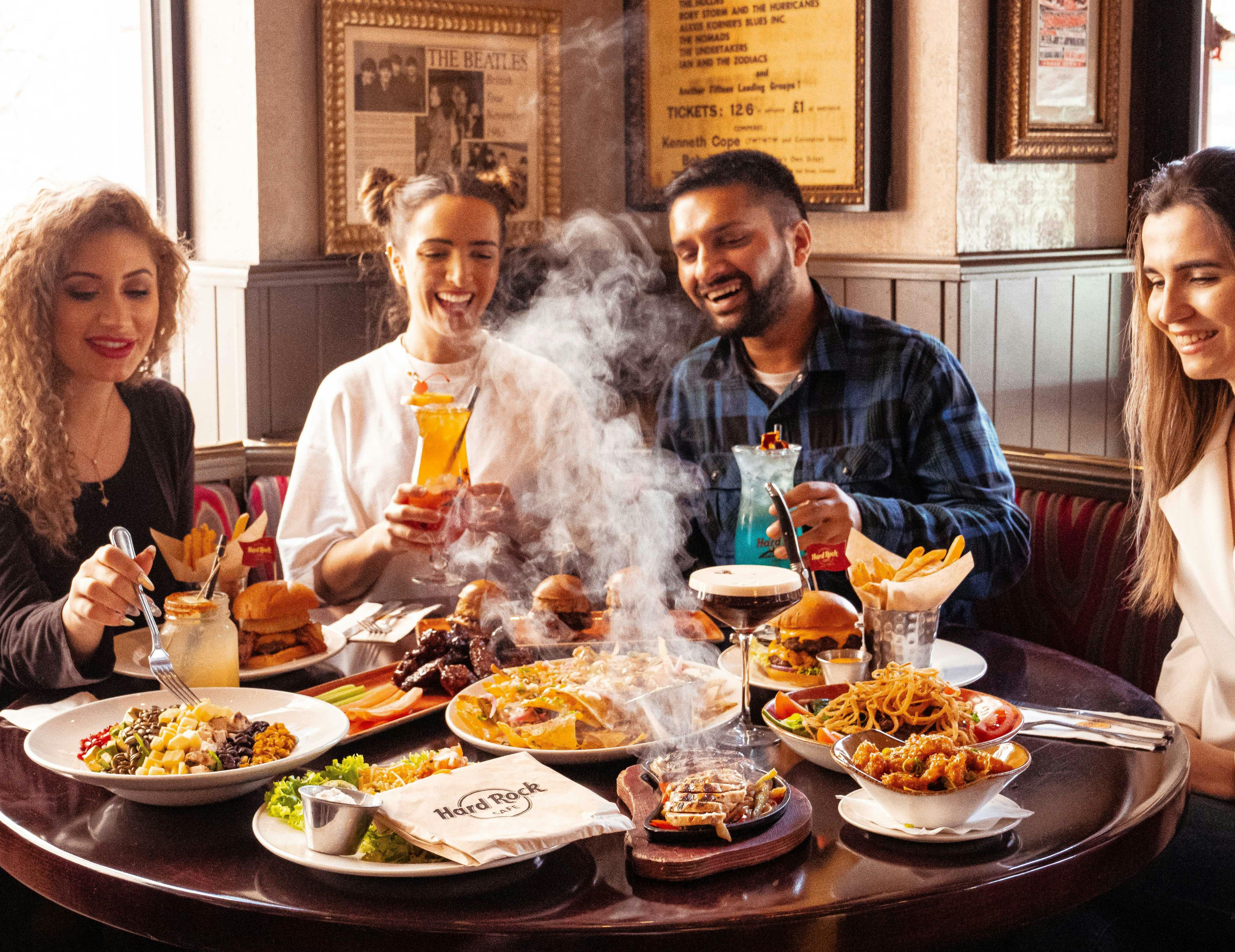 Three people are smiling and enjoying drinks and various dishes at a restaurant table, with steam rising from some of the food.