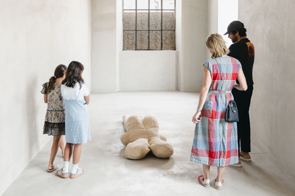 Four people are looking at a large soft toy bear lying on the floor in a brightly lit room, with a large window in the background.