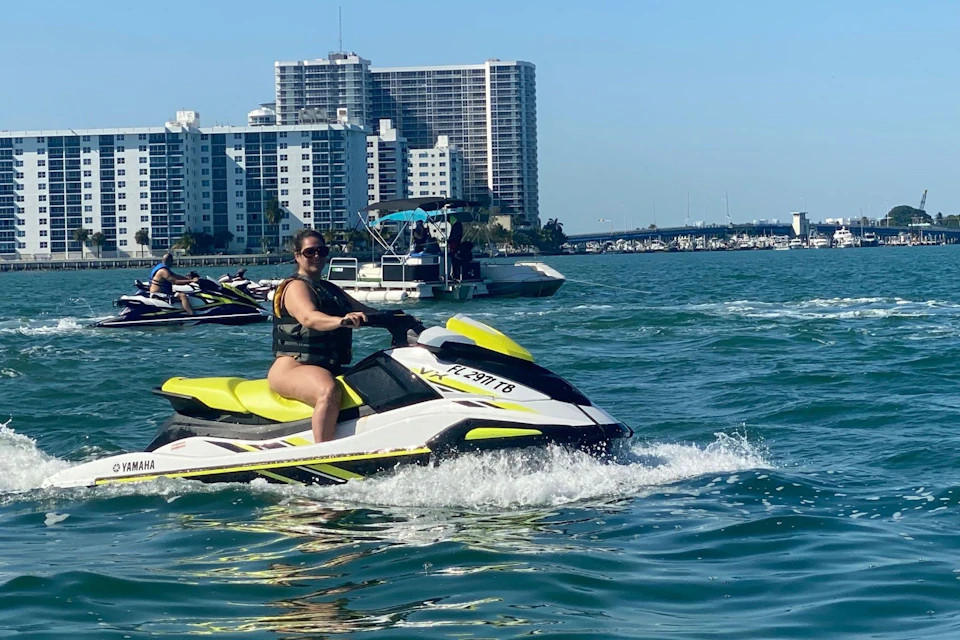 Person riding a jet ski on water, with boats and city buildings in the background under a clear blue sky.