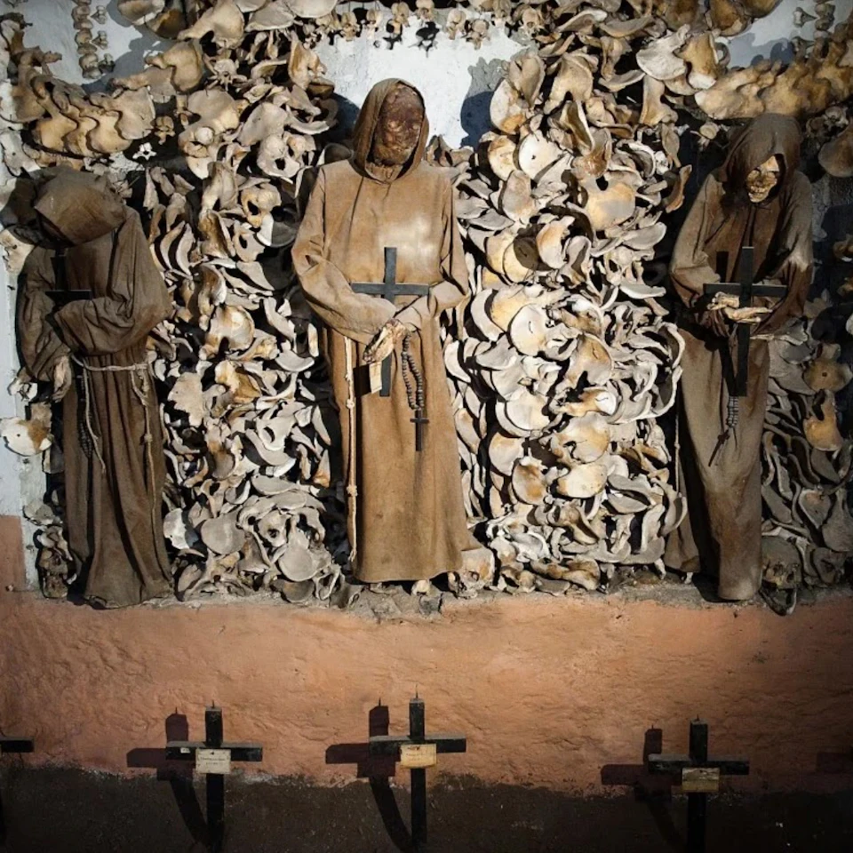 Three mummified monks in brown robes stand against a wall adorned with human bones, their hands holding crosses.