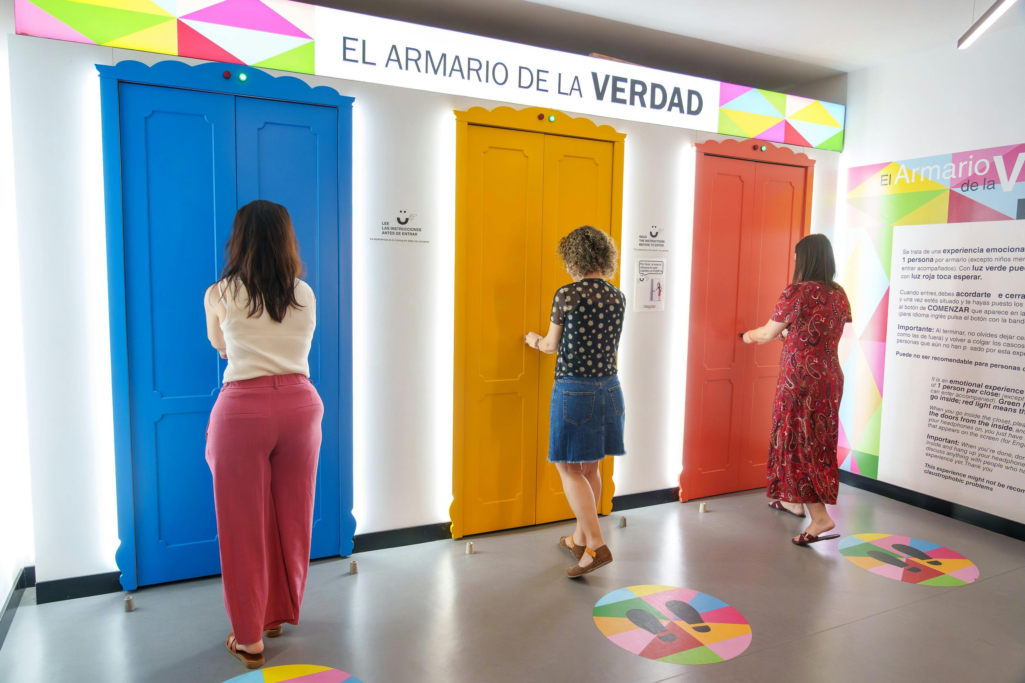 Três pessoas em pé em frente a portas coloridas com a inscrição «El Armario de la Verdad», numa sala bem iluminada.