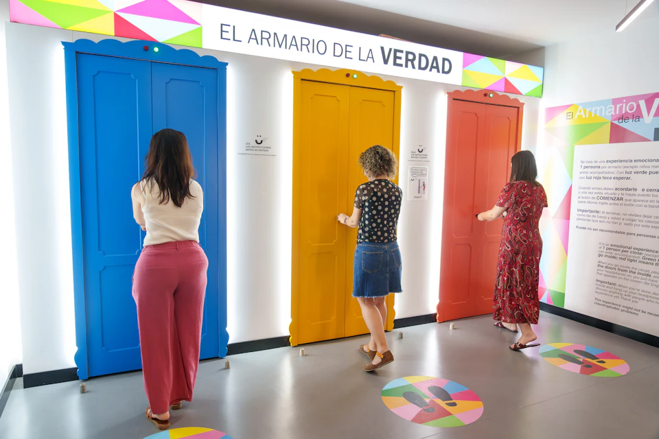Three people standing in front of colorful doors labeled "El Armario de la Verdad" in a brightly lit room.