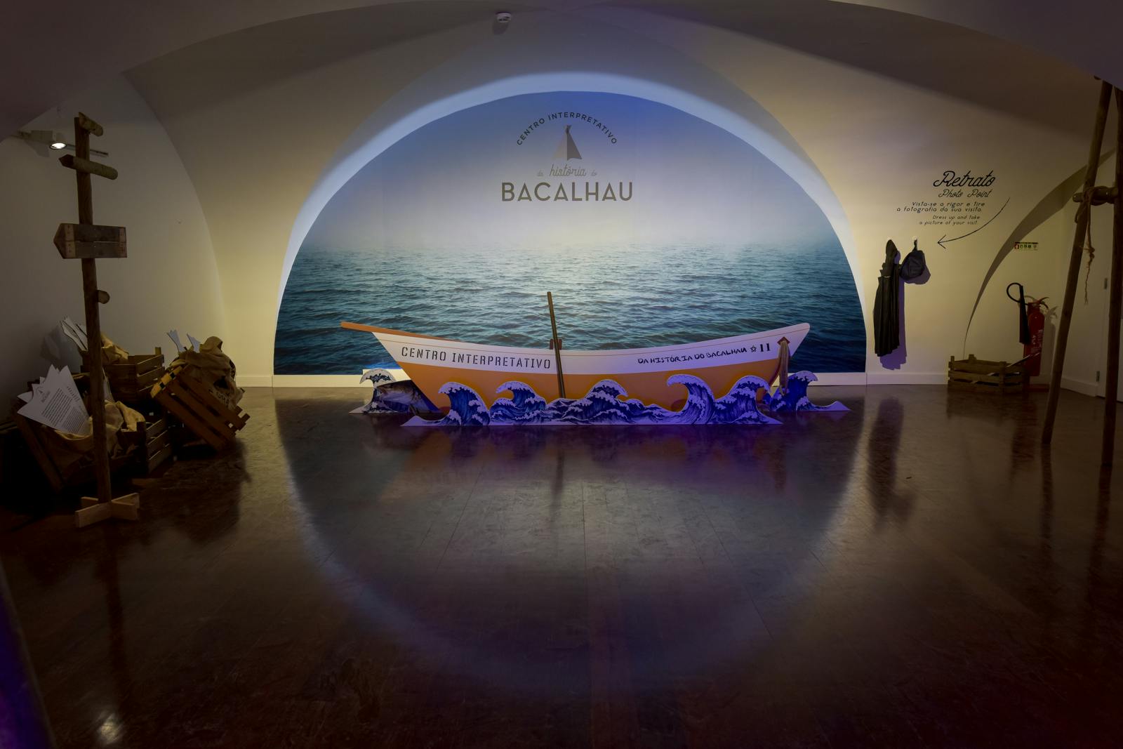 A small boat labeled &#34;Centro Interpretativo&#34; rests on a wave display in front of a wall mural of the sea with the text &#34;Bacalhau.&#34;