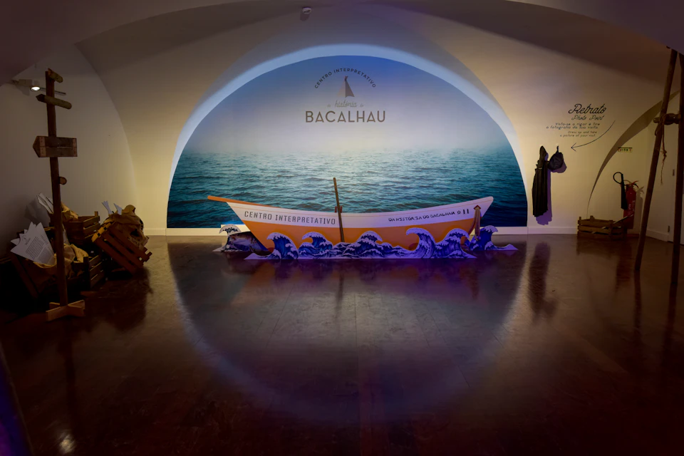 A small boat labeled "Centro Interpretativo" rests on a wave display in front of a wall mural of the sea with the text "Bacalhau."