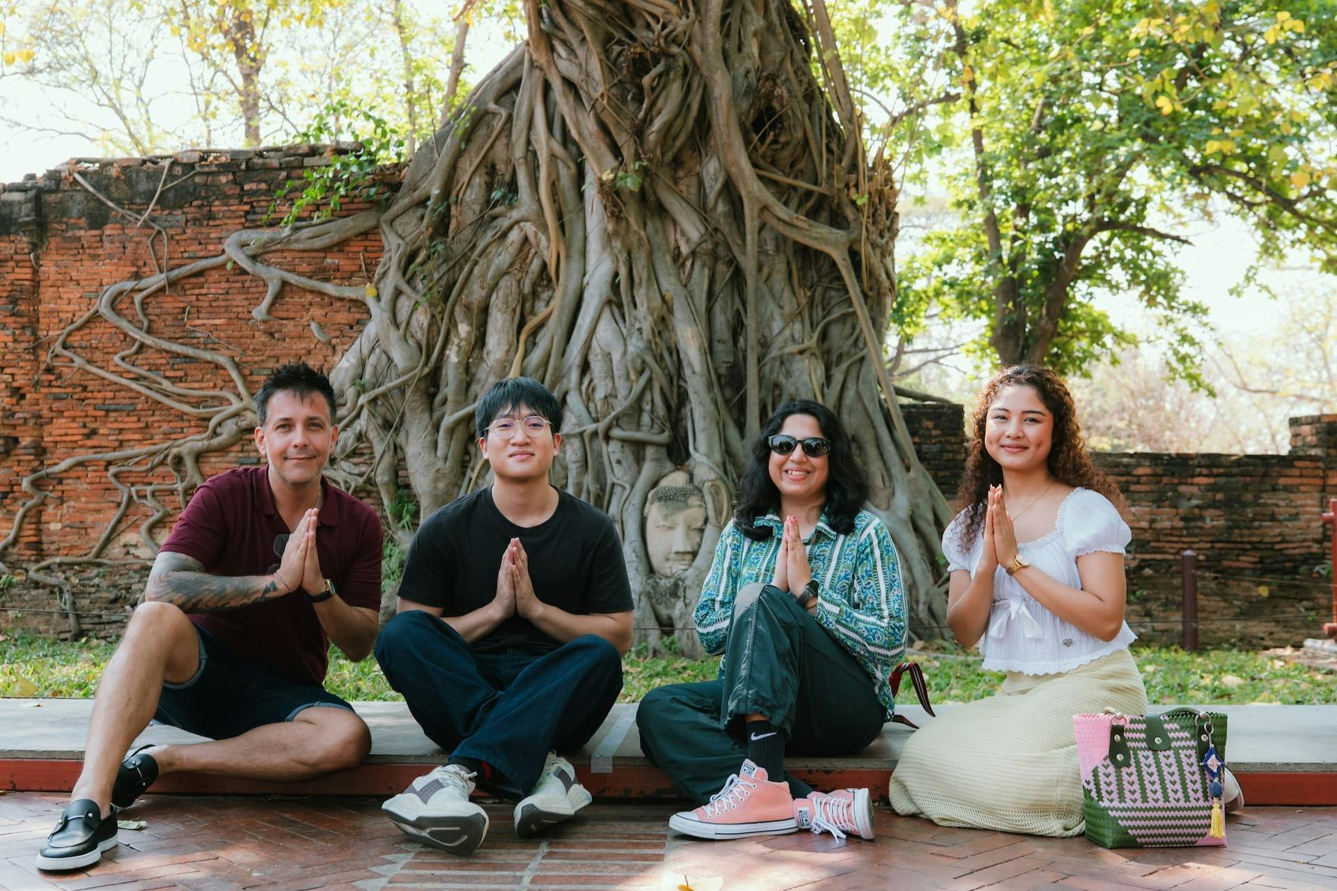 Tre personer sidder på jorden foran et træ med et Buddha-hoved indlejret i sine rødder og smiler og poserer med håndfladerne sammen.