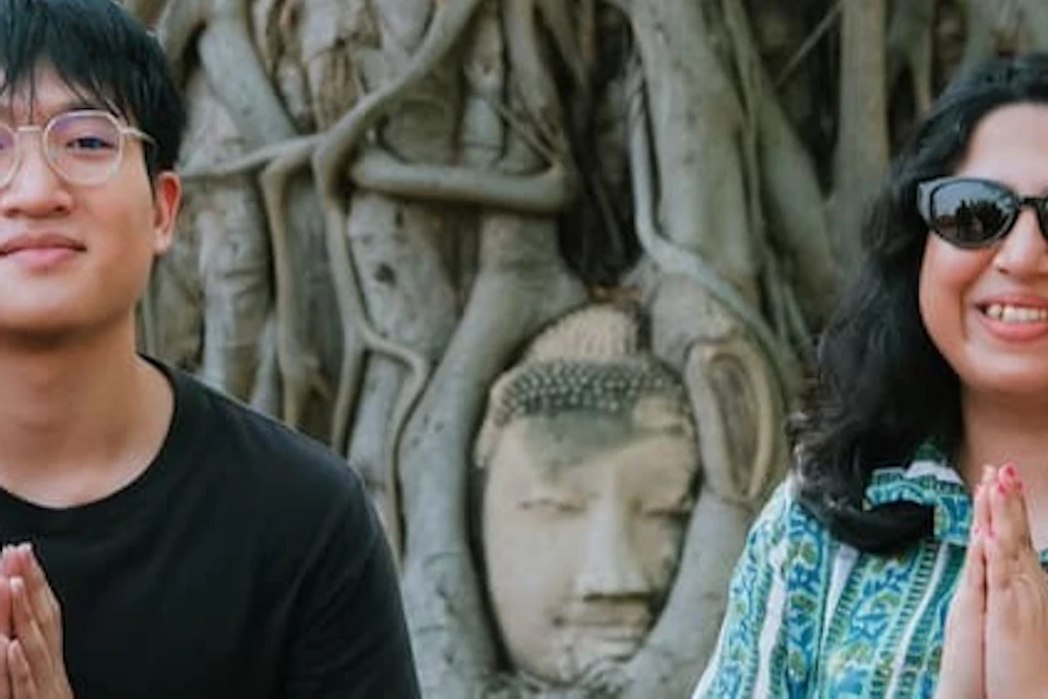 Three people sit on the ground in front of a tree with a Buddha head embedded in its roots, smiling and posing with palms together.