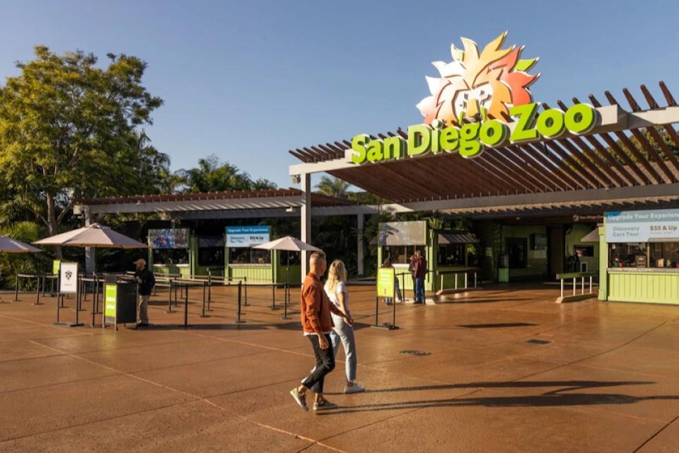 Two people walk past the entrance of the San Diego Zoo, which features a colorful lion logo and green sign under a clear sky.