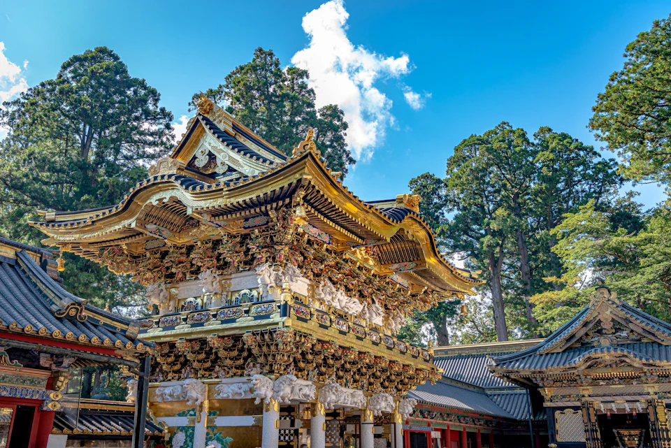 Elaborate traditional Japanese temple with intricate carvings and gold accents, surrounded by tall trees under a blue sky.