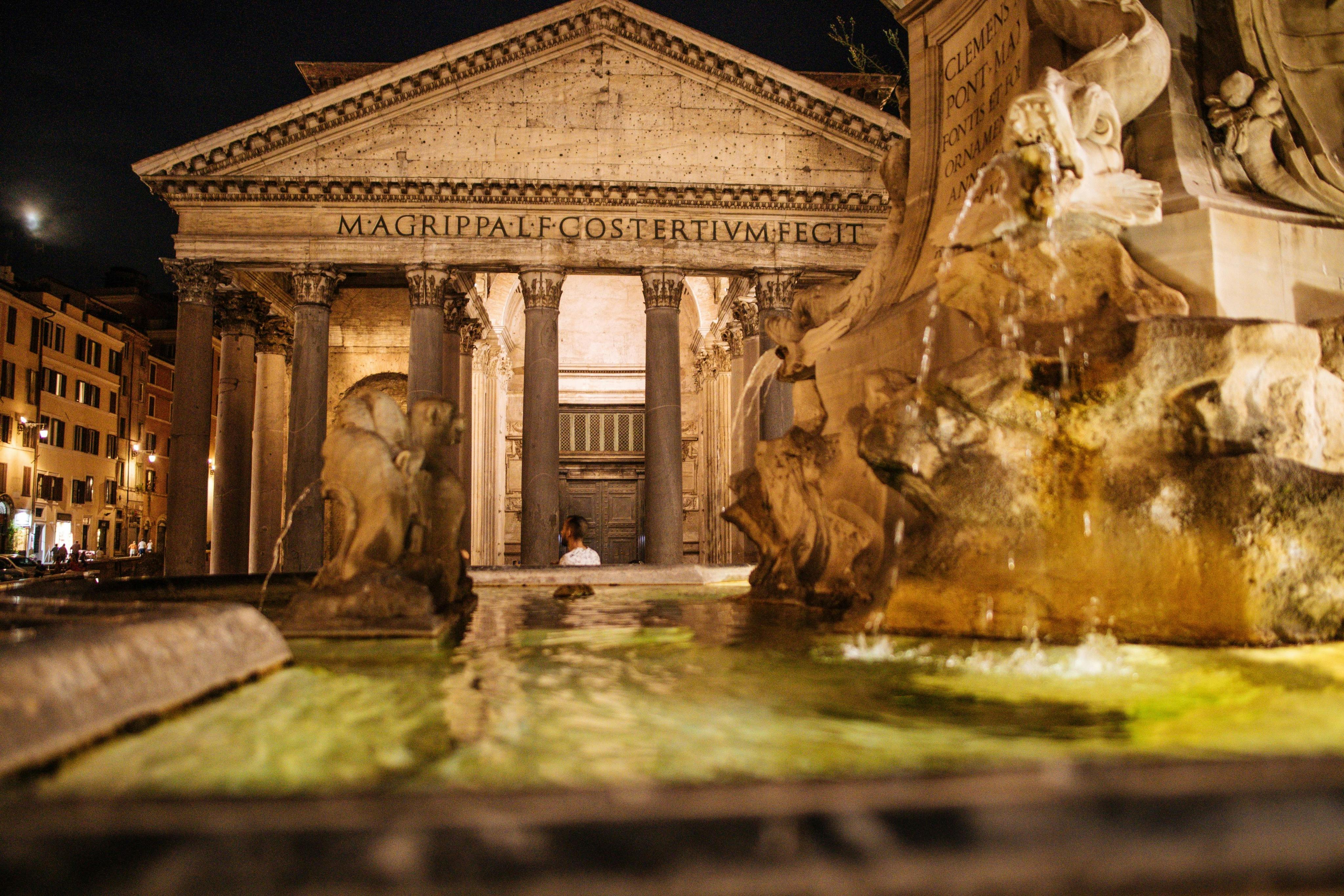 A nighttime view of the Pantheon in Rome with illuminated fountains in the foreground.