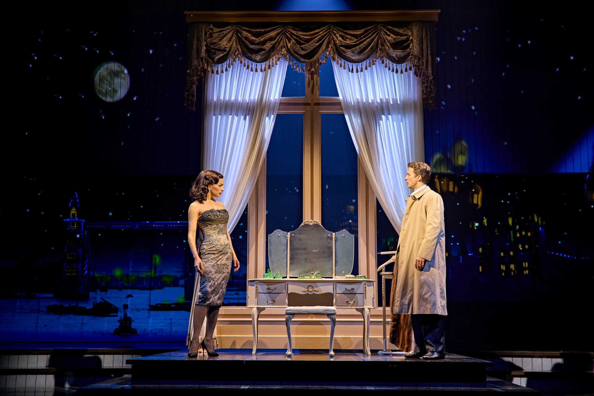 A theatrical scene with a woman in a dress and a man in a coat standing near a vanity, with a starry night backdrop.