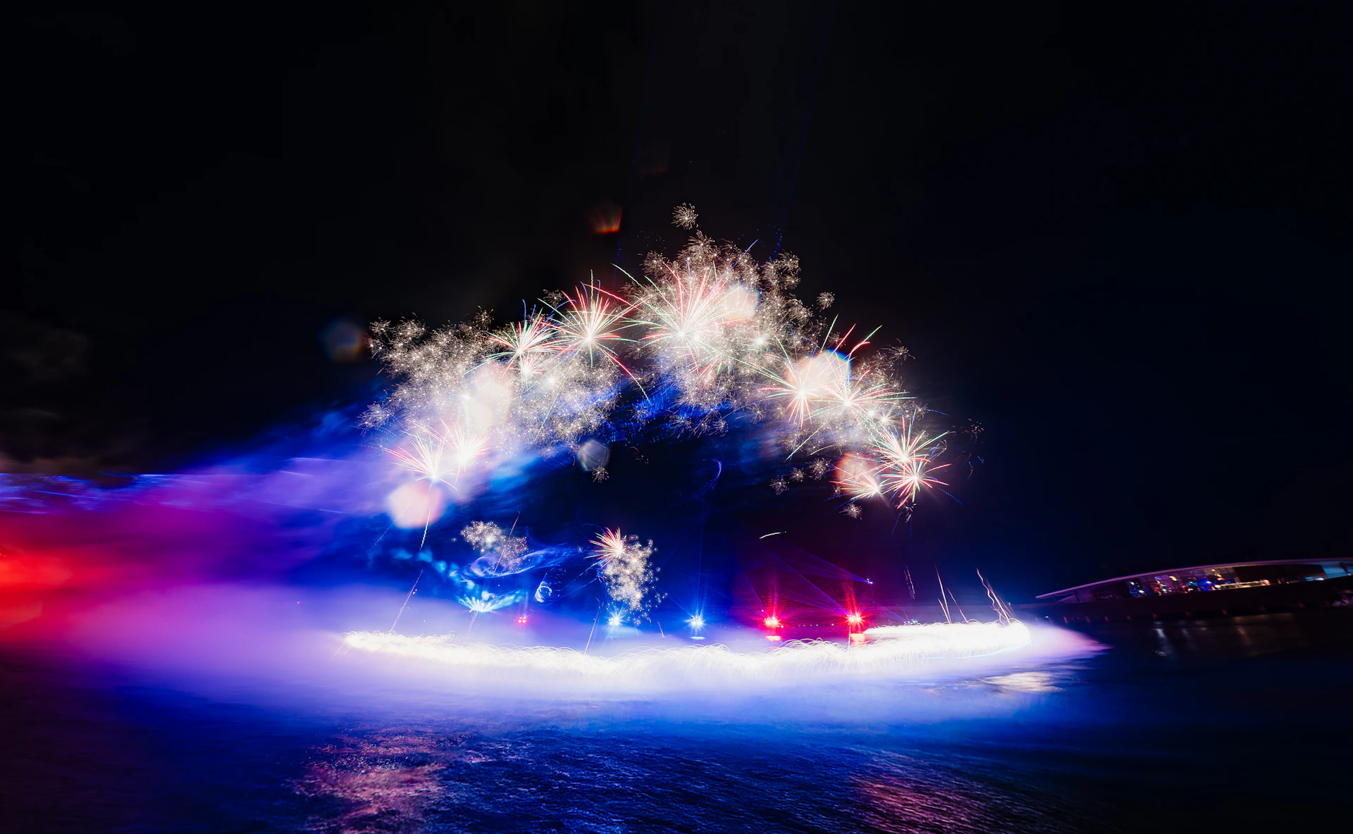 Fireworks bursting in the night sky above a misty scene illuminated by colorful lights, creating a festive atmosphere.