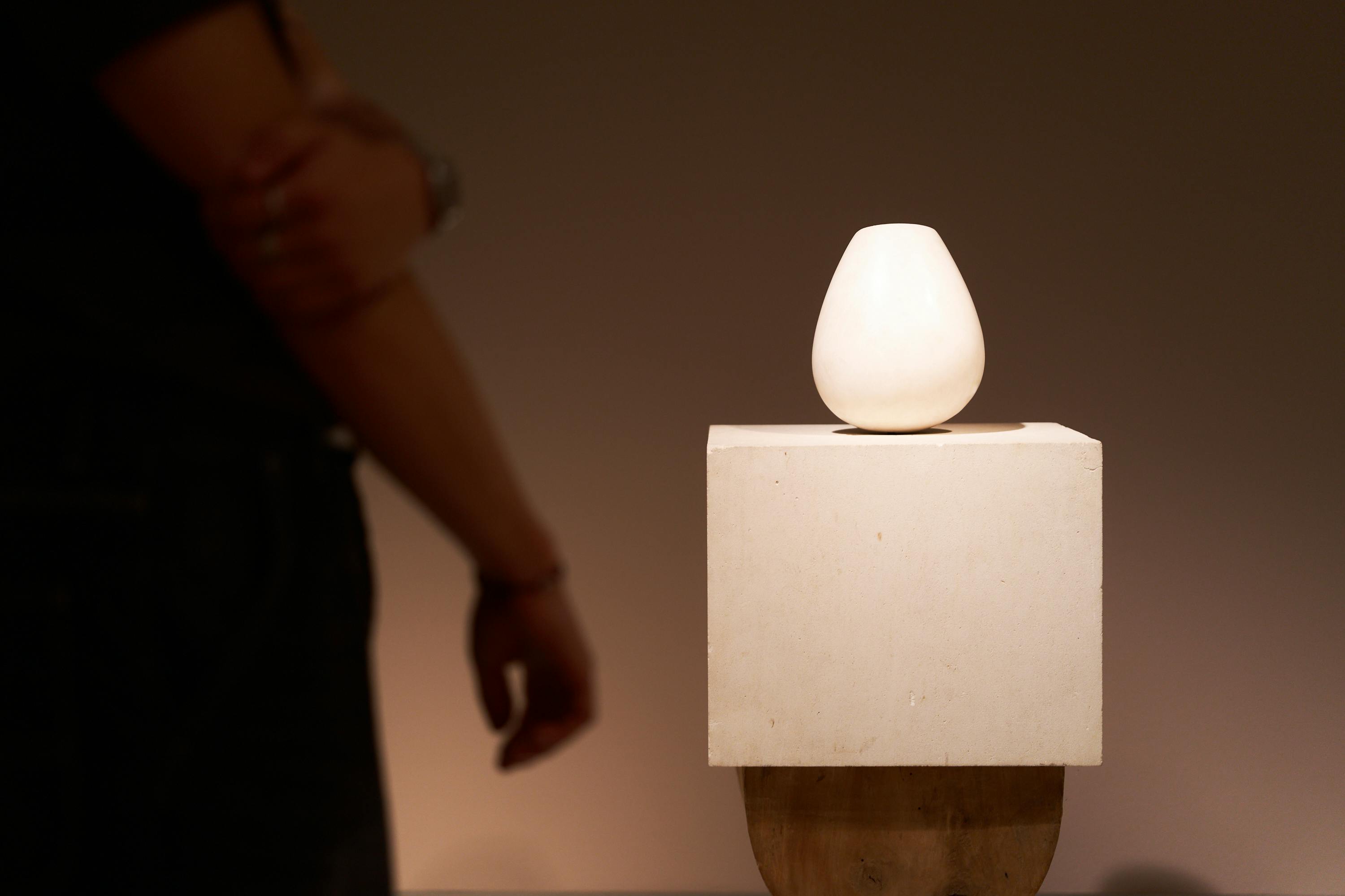 Un petit vase blanc est posé sur un socle carré. Une personne se tient à côté, les bras croisés, dans une pièce faiblement éclairée.
