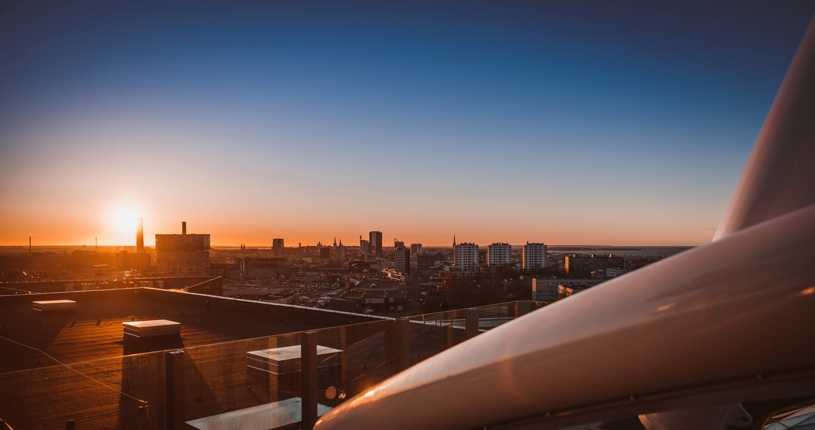 City skyline at sunset with tall buildings and rooftops, viewed from a balcony with clear glass railings.