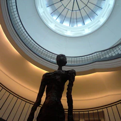 En mörk, abstrakt skulptur av en mänsklig figur står under ett cirkulärt takfönster i en byggnad med böjda, differentierade balkonger.