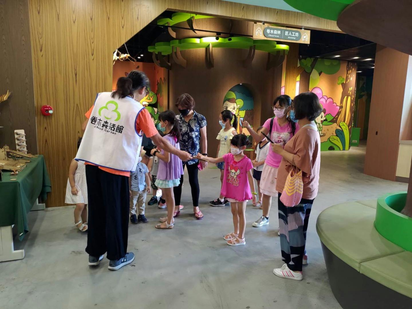 A group of adults and children interact inside a colorful, indoor play area with cartoon forest-themed decor.
