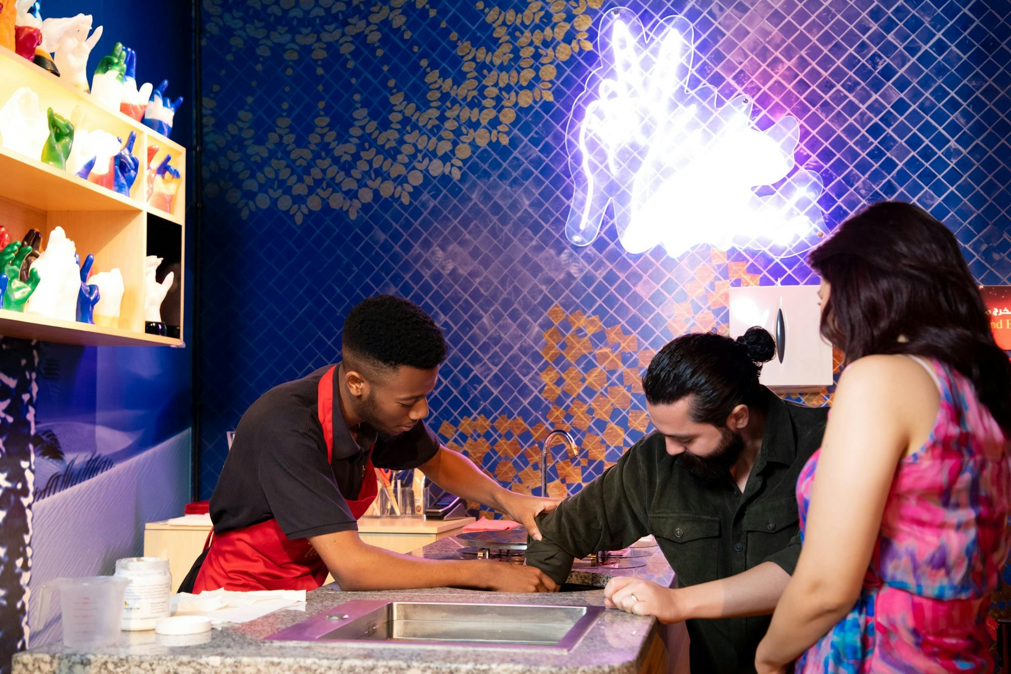 Ένας άνδρας με κόκκινη ποδιά βοηθά έναν άλλο άνδρα με ένα έργο μπροστά από έναν τοίχο με μπλε πλακάκια με μια λαμπερή απεικόνιση κουνελιού.