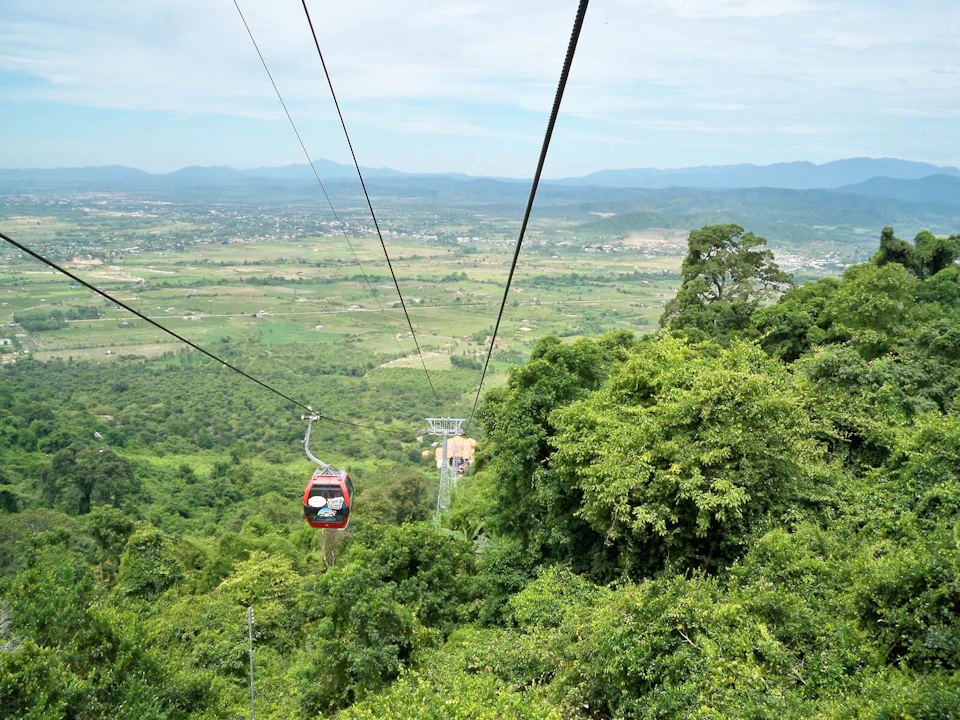 A red cable car traveling over lush green trees in a mountainous area with a valley and distant hills in the background.