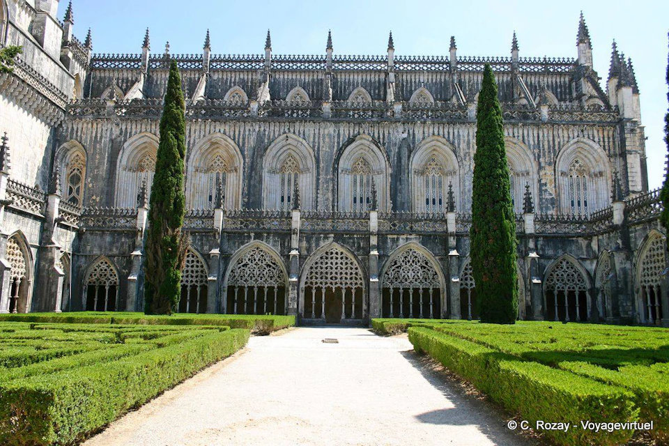 Gothic-style building with arched windows and intricate stone details, flanked by tall cypress trees and a manicured hedge garden.