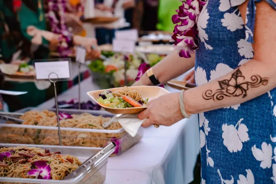 A person in a blue floral dress with a flower lei holds a plate of food at a buffet with trays of noodles and salad.