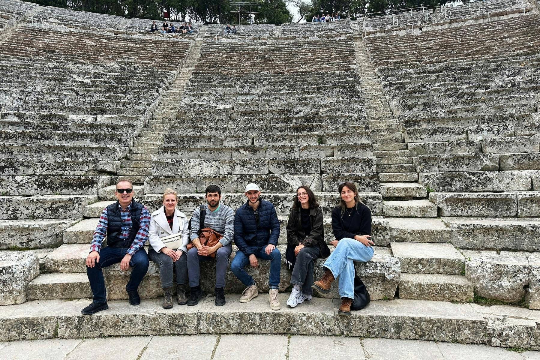 A group of people sit on the stone steps of the Epidaurus theater with others scattered above them.