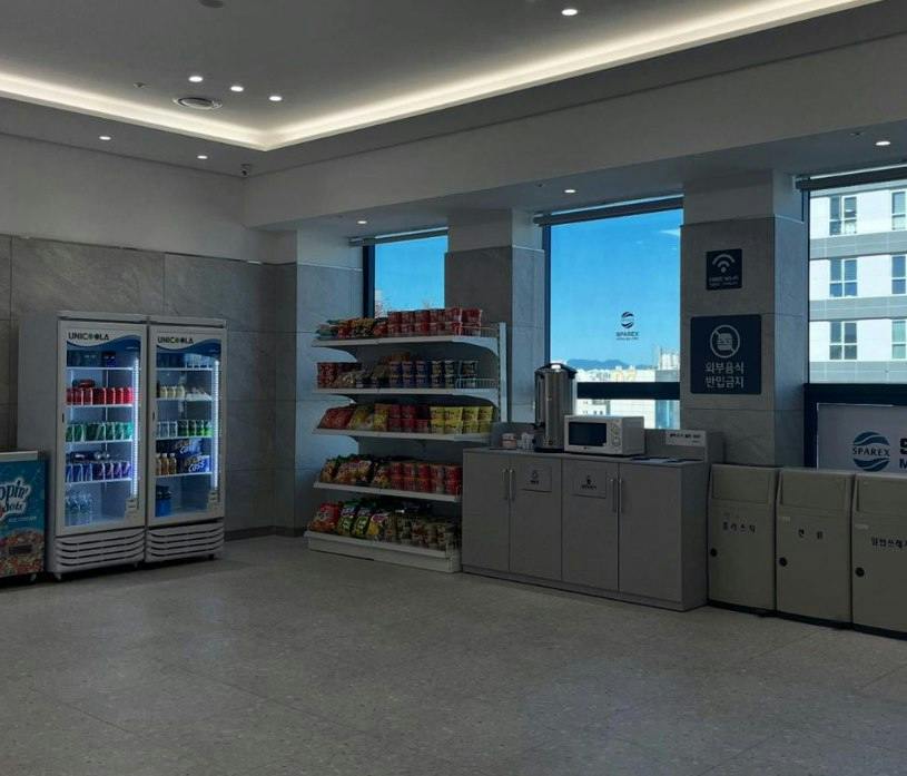 A break room with snack shelves, refrigerated drinks, a microwave, and a coffee dispenser by windows with a city view.