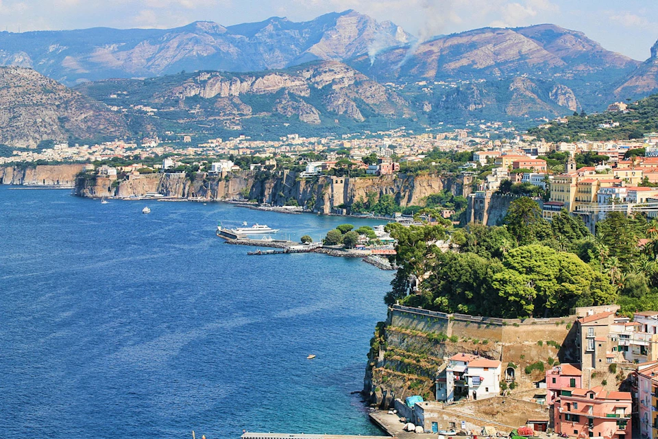 A coastal city with cliffs, buildings, and greenery overlooking a blue sea, backed by distant mountains under a partly cloudy sky.