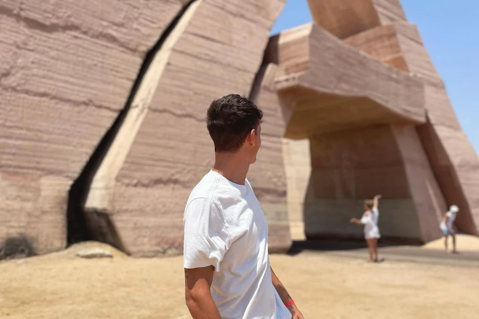 A person in a white shirt looks at large stone structures under a blue sky, with another person visible in the background.