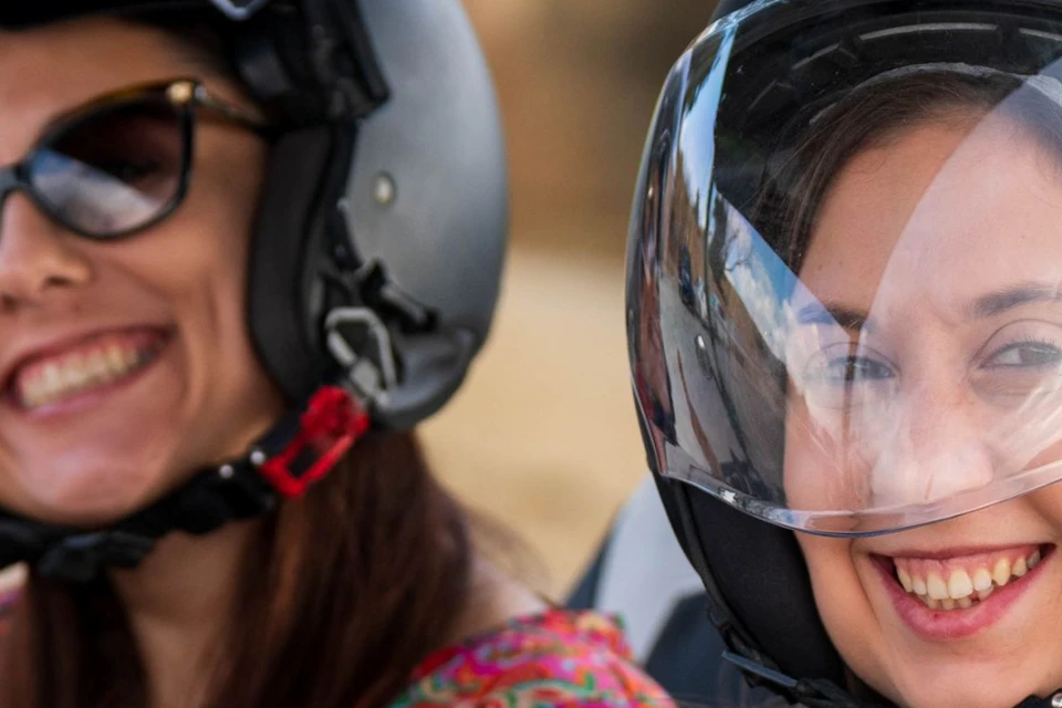 Two women wearing helmets and seated in a green-framed off-road vehicle, smiling and gesturing thumbs-up and peace sign.