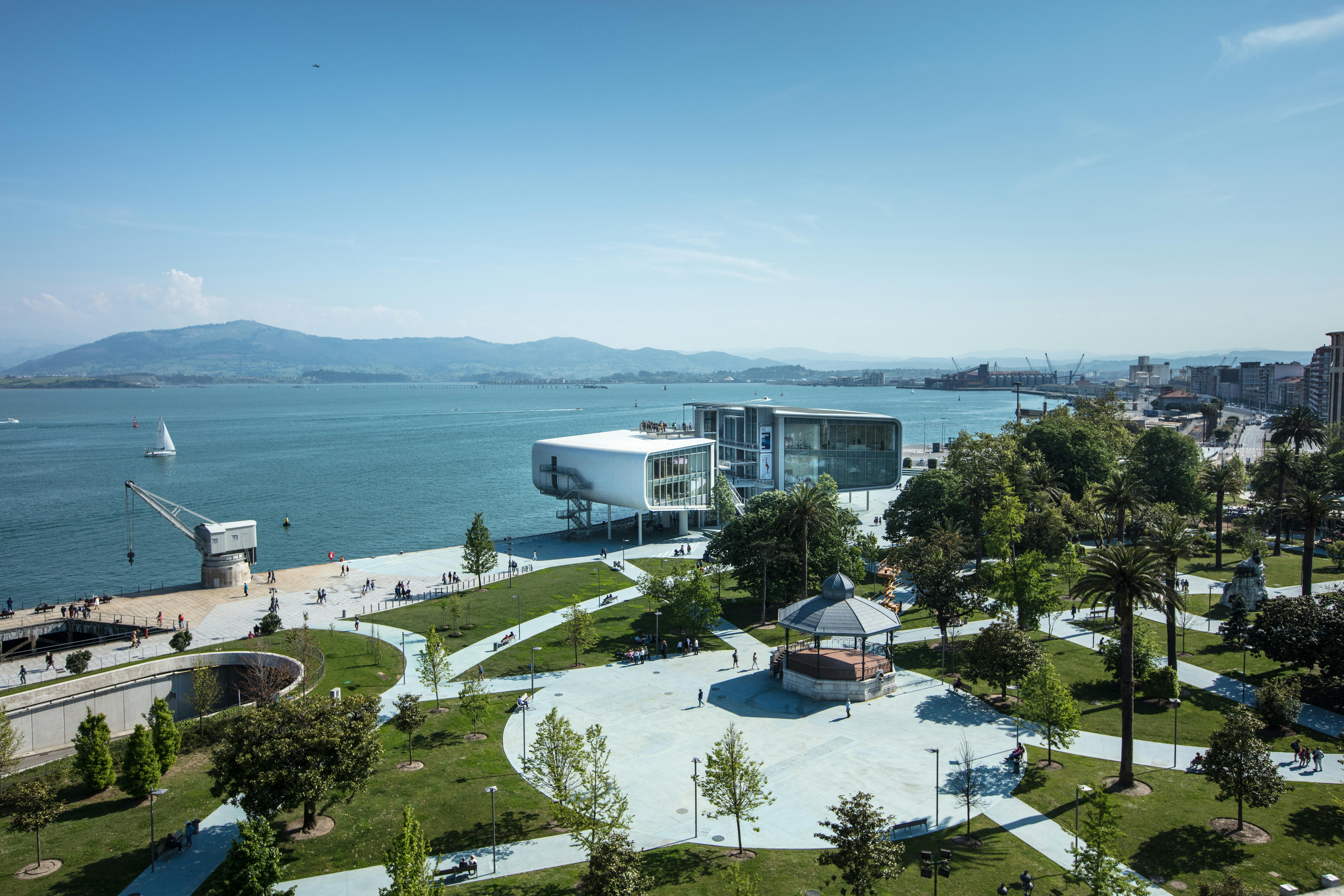 Ένα σύγχρονο παραθαλάσσιο κτίριο έχει θέα σε ένα πάρκο με δέντρα, μονοπάτια και κιόσκι. Το σκηνικό τοποθετείται με φόντο τα βουνά και το νερό.