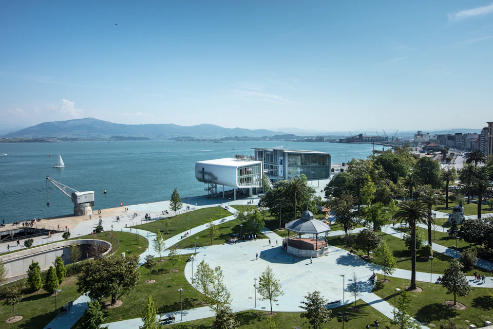 A modern waterfront building overlooks a park with trees, pathways, and a gazebo. The scene is set against a backdrop of mountains and water.