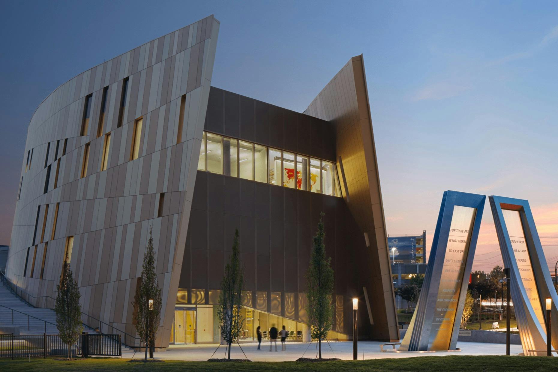 Um edifício moderno com uma arquitetura angular e grandes janelas, iluminado ao anoitecer, com árvores e um monumento em primeiro plano.