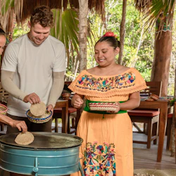 A man and a woman learn to make tortillas from a woman in traditional dress at an outdoor cooking station with wooden tables nearby.