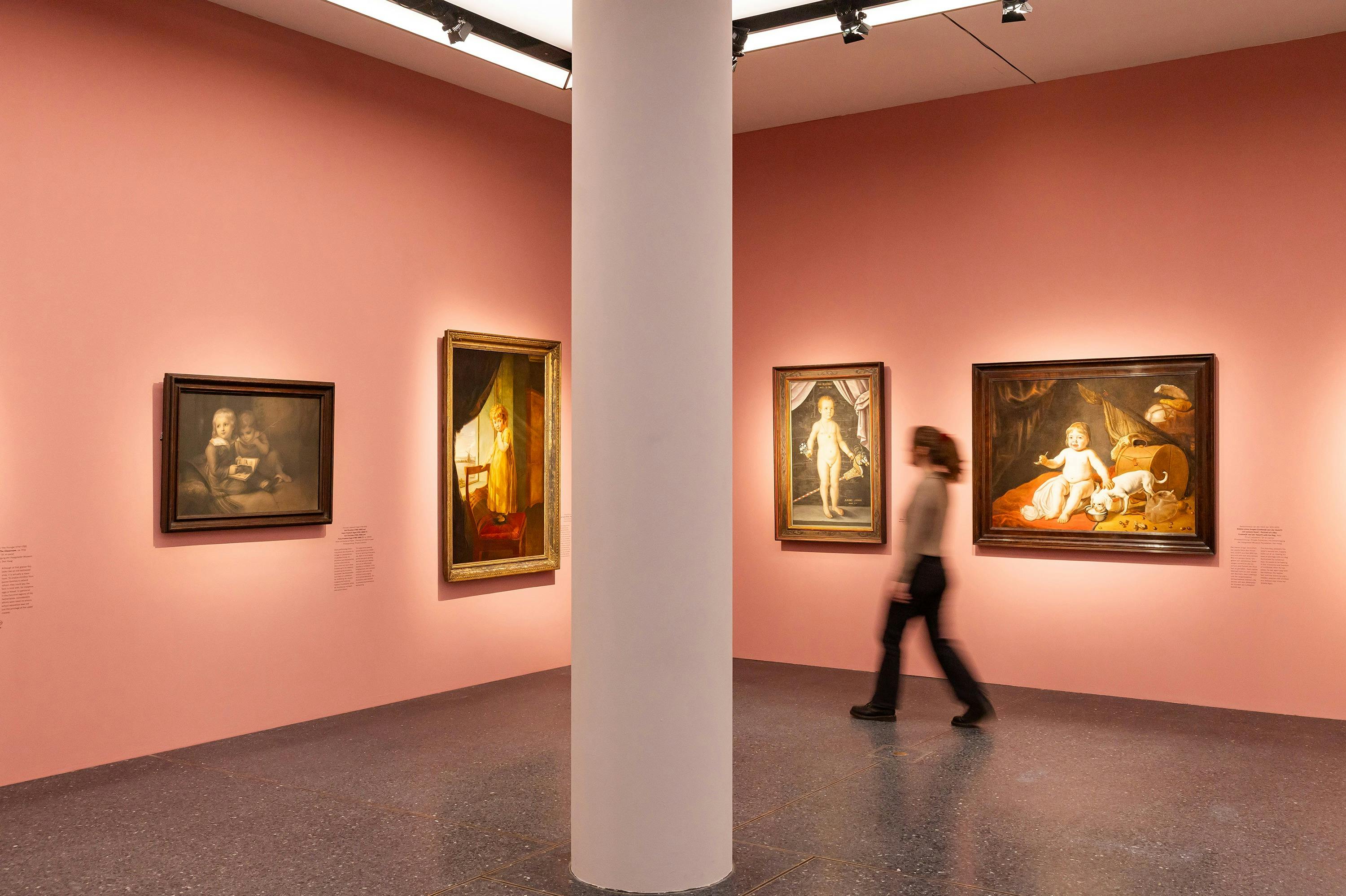 분홍색 벽에 그림이 그려진 미술관, 중앙에 기둥이 있고 미술품을 지나가는 사람이 있습니다.