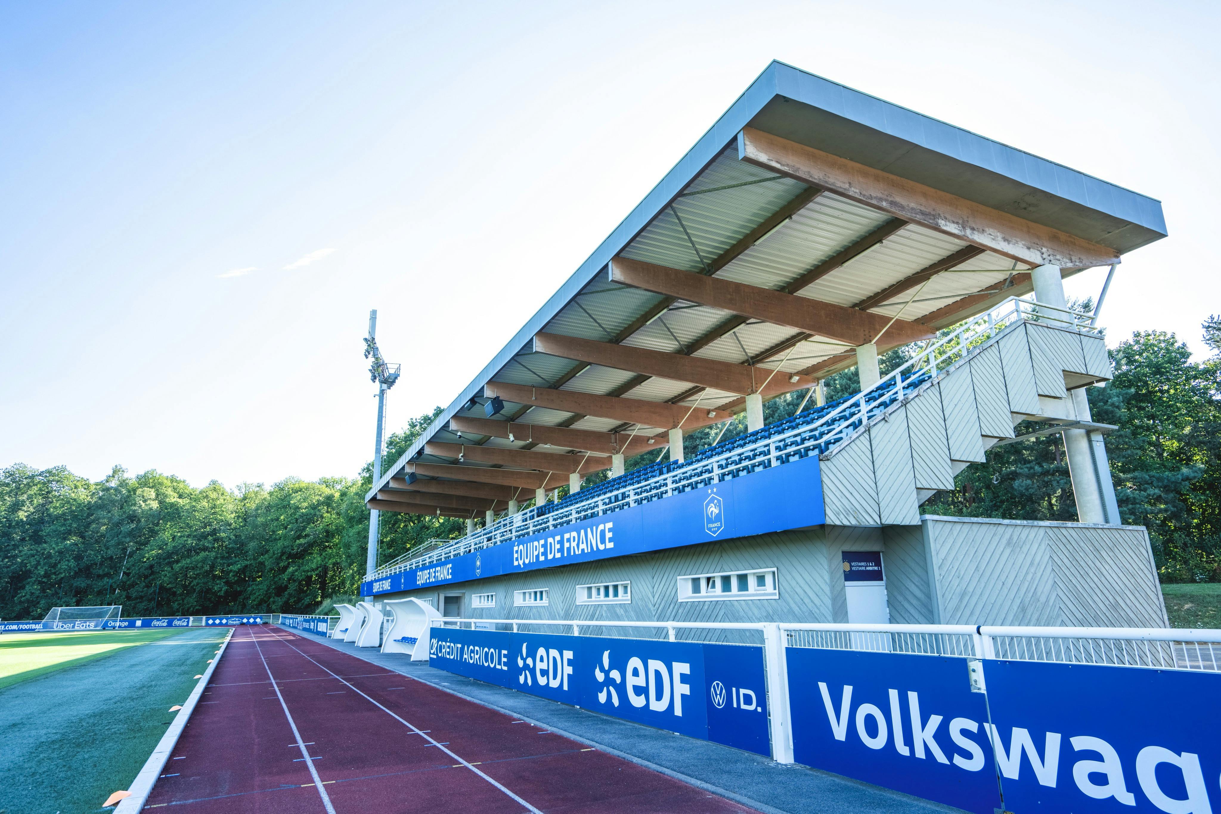 En liten fotbollsstadion med sittplatser, en röd löpbana och skogsklädda omgivningar under en klar himmel.