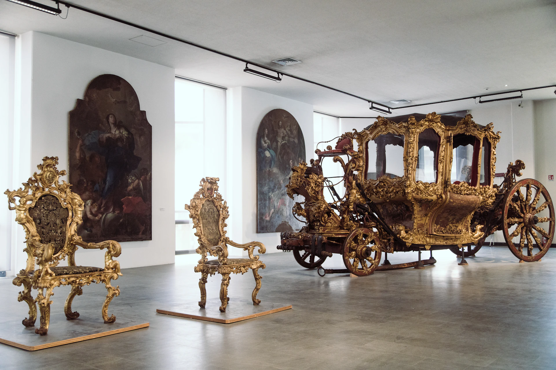 Museum exhibit featuring an ornate gold carriage and two intricately carved chairs, with large framed paintings on the walls behind them.