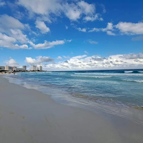 Ein ruhiger Strand mit sanften Wellen, ein blauer Himmel mit vereinzelten Wolken und Hochhäuser in der Ferne. Nur wenige Menschen gehen spazieren.